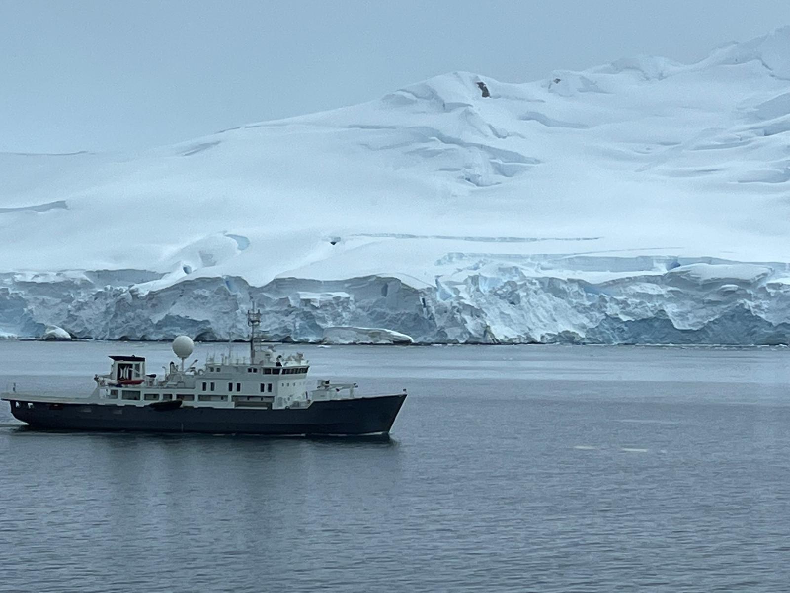 A small expedition cruise ship sails through calm Antarctic waters, framed by snow-covered glaciers and ice-covered mountains.