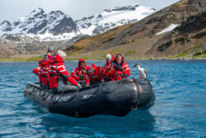 Luxury expedition cruise guests explore Antarctica by Zodiac, surrounded by crystal-blue waters, towering snow-capped mountains, and an up-close encounter with a penguin.
